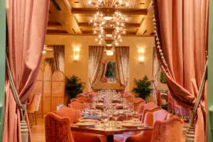Main Dining Room with pink velvet chairs, chandeliers, and floral drapes, viewed through parted rose curtains.