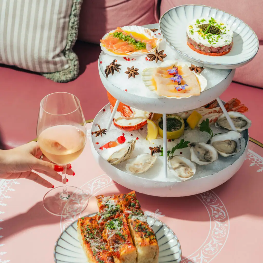 Seafood tower with oysters, shrimp, and crudo on ice, served with focaccia and a glass of rosé at Casadonna Miami, on a blush-pink patio setting.