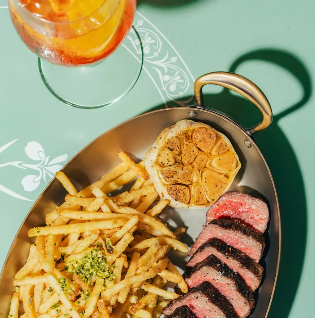 Top-down view of steak frites served with roasted garlic and shoestring fries, paired with an orange spritz cocktail at Casadonna Miami.
